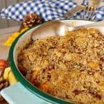 A close-up of sweet potato casserole, topped with a golden, crumbly pecan mixture, baked to a crisp. The spoon beside the dish suggests it's ready to serve, with autumnal accents and a cozy kitchen vibe in the background.
