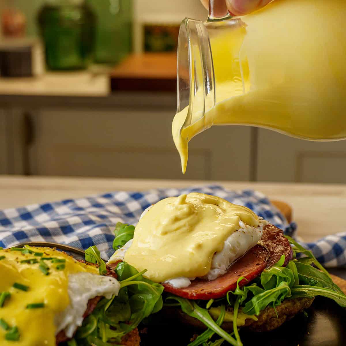 A plate of Eggs Benedict topped with hollandaise sauce, with the sauce being poured over one poached egg, surrounded by vibrant greens and set on a black plate with a checkered napkin in the background.