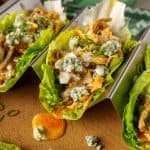 Buffalo chicken lettuce wraps displayed on a wooden board, garnished with blue cheese, green onions, and a bottle of hot sauce in the background.