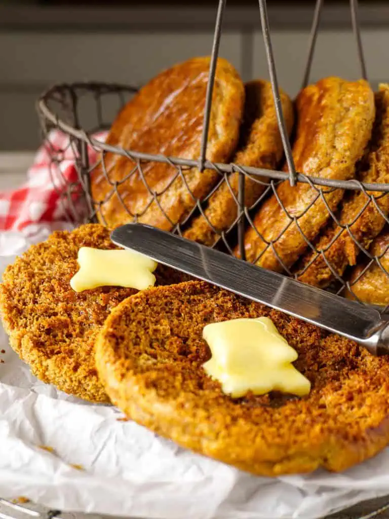 A warm and inviting scene showcasing a wire basket filled with keto English muffins, with a focus on a toasted muffin half smeared with melting butter on parchment paper.