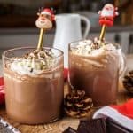 Two mugs of keto hot chocolate on a wooden surface, garnished with whipped cream, chocolate shavings, and festive decorations, including pinecones and dark chocolate squares.