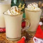 Close-up of two festive glasses of sugar-free eggnog topped with whipped cream, garnished with golden antler stirrers, placed on a wooden board surrounded by a Santa hat and holiday decorations.