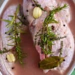 Top-down view of turkey breast in brine, garnished with fresh rosemary, thyme, garlic cloves, and black peppercorns.