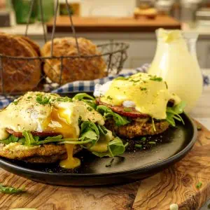 Keto eggs Benedict served on a black plate, with hollandaise sauce, greens, and bacon, set against a rustic kitchen backdrop.