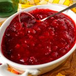 White serving dish filled with sugar-free cranberry sauce on a wooden board, surrounded by autumn leaves and fresh cranberries.