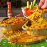 A spoon lifting a portion of Game Day Dip from a dish, showcasing creamy, melted cheese and a golden crust, with celery and bell pepper sticks blurred in the background.