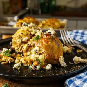 A plate of golden-brown stuffed chicken thighs topped with feta cheese and fresh herbs, served with a fork and a blue checkered napkin in the background.