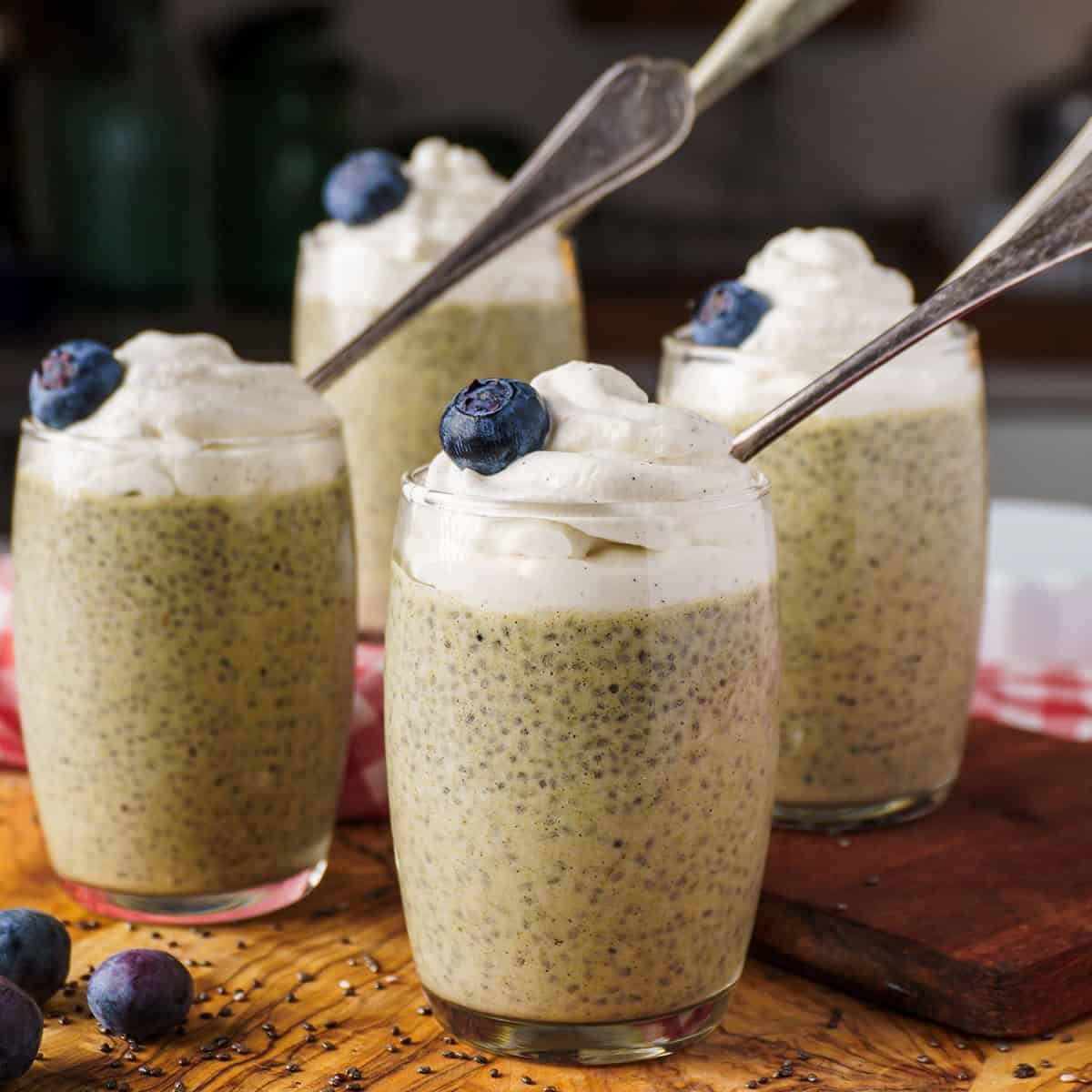 Chia_Seed_Protein_Pudding_featured_image Four glasses of chia seed protein pudding topped with whipped cream and blueberries, arranged on a wooden surface with vintage spoons.