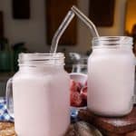 A cold coconut smoothie in a mason jar, set on a rustic wooden board with a bowl of frozen strawberries and a checkered napkin in the background.