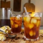 Two glasses of cold brew coffee with ice and glass straws, with a pitcher of coffee and a small jug of cream in the background.