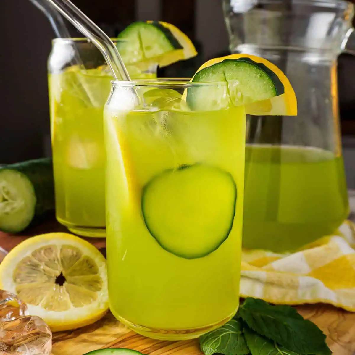 Cucumber_Lemonade_featured_image Cucumber lemonade in a glass pitcher with glasses filled with ice, lemon, and cucumber slices, placed on a wooden board with fresh ingredients.