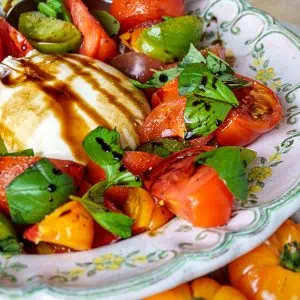 Colorful heirloom tomatoes and burrata with balsamic glaze and basil in a green and yellow rimmed vintage plate, photographed from the side.