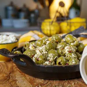 Cast iron skillet filled with green olives and crumbled cheese, set on a wood surface with a yellow checkered cloth and a small bowl of cheese in the background.