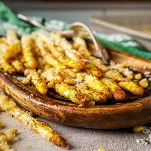 Roasted white asparagus arranged on a wooden dish, garnished with pine nuts and cheese, placed on a rustic tabletop.
