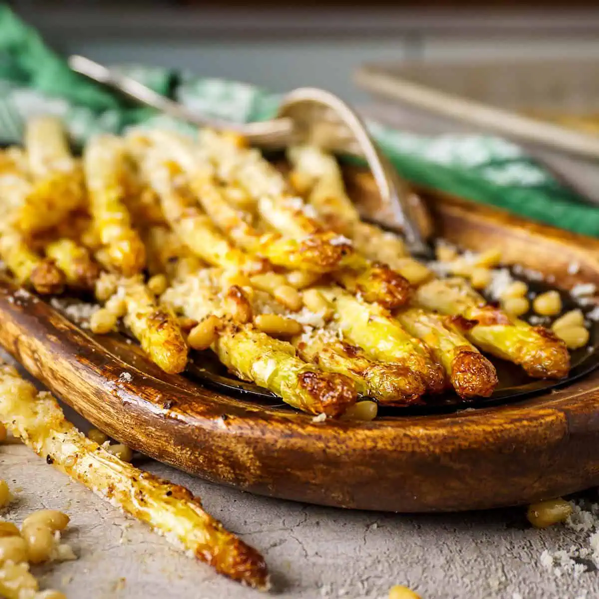 Roasted white asparagus arranged on a wooden dish, garnished with pine nuts and cheese, placed on a rustic tabletop.